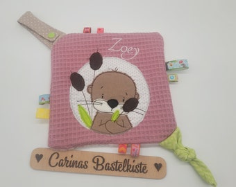 Puede incluir: Un juguete para bebés cuadrado rosa con textura de gofre, con un diseño de nutria bordada. La nutria está rodeada por un círculo blanco con juncos marrones. El nombre "Zoey" está bordado encima de la nutria. El juguete tiene cintas coloridas y una tela verde anudada.