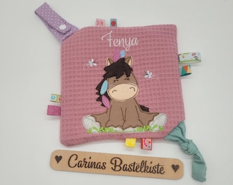 Puede incluir: Un juguete sensorial para bebés rosa texturizado con una aplicación de caballo de dibujos animados. El juguete tiene un cierre de botón, cintas de colores y el nombre "Fenya" bordado. También hay una etiqueta de madera con el texto "Carinas Bastelkiste".