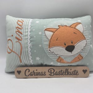 Könnte beinhalten: Ein rechteckiges Kissen aus hellgrünem Stoff mit weißen Sternen und einem gestreiften Bereich. Der Name Lina ist in orangefarbener Schreibschrift gestickt. Ein zentraler runder Aufnäher zeigt einen Fuchs. Ein Holzschild trägt die Aufschrift Carinas Bastelkiste.