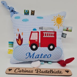Könnte beinhalten: Ein handgemachtes, quadratisches, blau-weiß kariertes Kissen mit einem roten, gestickten Feuerwehrauto-Design. Das Kissen hat den Namen "Mateo" in blauer Schrift, eine gelbe Sonne und verschiedene bunte Etiketten. Das Holzschild darunter lautet "Carinas Bastelkiste".