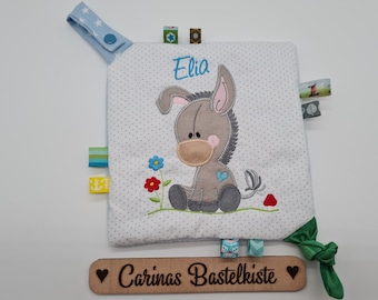 Puede incluir: Un cuadrado sensorial para bebés con un aplique de burro gris, el nombre "Elia" bordado en azul y cintas de colores. El cuadrado es blanco con lunares grises y tiene una pestaña azul con estampado de estrellas. Un letrero de madera dice "Carinas Bastelkiste".