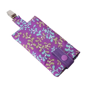 Könnte beinhalten: Lilafarbener Stoffbeutel mit einem Blumenmuster in Grün- und Blautönen. Der Beutel hat einen Clip oben und einen lilafarbenen Knopfverschluss.