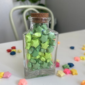 May include: A glass jar filled with green origami stars, with a cork stopper. The jar is sitting on a white surface with scattered origami stars in various colors.