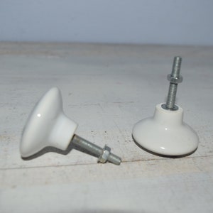 May include: Two white ceramic cabinet knobs with silver metal screws. The knobs are round with a slightly raised center and a flat base.