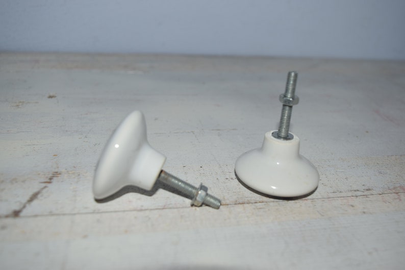 May include: Two white ceramic cabinet knobs with silver metal screws. The knobs are round with a slightly raised center and a flat base.