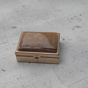 Op de afbeelding: Een klein, vierkant, goudkleurig metalen doosje met een scharnierend deksel. Het deksel is gemaakt van een doorschijnend, lichtbruin materiaal met een marmerpatroon.