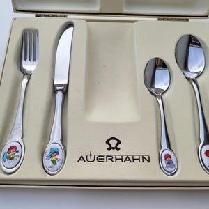 Könnte beinhalten: Auerhahn Kinderbesteck-Set in einer cremefarbenen Box. Das Set enthält eine Gabel, ein Messer und zwei Löffel. Jedes Teil hat einen silbernen Griff mit einem farbenfrohen Cartoon-Charakter-Design. Die Box ist geöffnet.