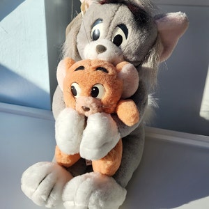 May include: A plush toy of Tom Cat from Tom and Jerry, holding a smaller plush toy of Jerry Mouse. Tom is gray with black eyes and a black nose. Jerry is orange with black eyes and a black nose.
