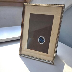 May include: A rectangular silver-toned metal picture frame with a cream-coloured matting. The frame contains a dark brown backing board featuring a circular sticker with a blue and black logo that displays the text JACIE in a stylised font.
