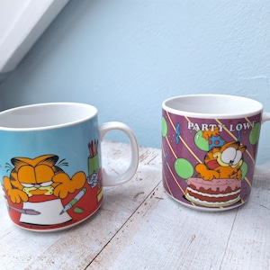 Könnte beinhalten: Zwei Keramikbecher mit Garfield der Katze. Ein Becher hat einen blauen Hintergrund mit Garfield, der an einem Schreibtisch schreibt. Der andere Becher hat einen lila Hintergrund mit Garfield, der eine Partymütze trägt und einen Kuchen hält. Der Text auf dem Becher lautet "Party Lowe".