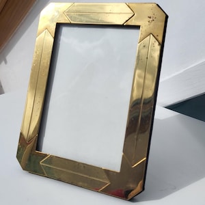 May include: A rectangular picture frame with a polished gold-tone metal border. The frame features a geometric design with angled corner segments and straight side pieces. The surface of the metal shows minor signs of age and light tarnishing.