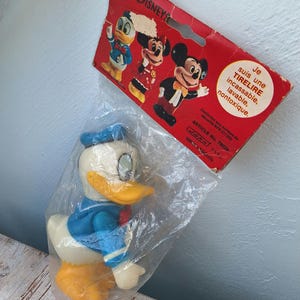 Può includere: Un giocattolo vintage di plastica di Paperino con una camicia da marinaio blu e pantaloni gialli. Il giocattolo è confezionato in un sacchetto di plastica trasparente con un retro in cartone rosso e bianco che dice "WALT DISNEYS" e "Je suis une TIRELIRE incassable, lavable, nontoxique."