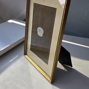 Puede incluir: Un marco de fotos rectangular con un borde de metal fino en tono dorado y un borde texturizado. El marco cuenta con un paspartú de color crema que rodea una inserción de tela marrón oscuro. Una pequeña pegatina ovalada blanca con el texto ROJA Versilber está centrada.