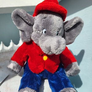 Könnte beinhalten: Ein Stofftier-Elefant mit grauem Körper, großen Ohren und einem Rüssel. Er trägt eine rote Jacke, blaue Hose und einen roten Hut. Die Jacke hat einen gelben Knopf. Das Spielzeug ist als Cartoon-Figur gestaltet.