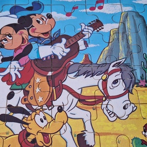 Può includere: Un puzzle vintage con Mickey Mouse, Minnie Mouse e Pluto vestiti da cowboy. Mickey suona la chitarra mentre cavalca un cavallo bianco con Minnie. Pluto corre accanto al cavallo. Il puzzle è ambientato in un paesaggio desertico con cactus e una montagna sullo sfondo.