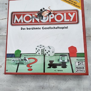 Op de afbeelding: Een rood, wit en blauwe Monopoly-speldoos met de naam van het spel in grote witte letters. De doos heeft een cartoonafbeelding van een man met een hoge hoed en monokel die een stok vasthoudt. De tekst "Das berühmte Gesellschaftsspiel" staat onder de naam van het spel. De doos bevat ook een klein beeld van een auto en dobbelstenen.