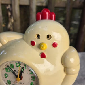 May include: A cream-colored chicken-shaped analog clock with a red comb, red cheeks, and a yellow beak. The clock face features green numbers from 1 to 12, black hour and minute hands, and a yellow second hand with the text "Quartz" printed below.