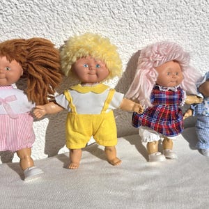 Könnte beinhalten: Eine Sammlung von fünf Puppen mit unterschiedlichen Merkmalen. Die erste hat braune Wollhaare und ein rosa gestreiftes Kleid. Die zweite hat gelbes Haar und eine gelbe Latzhose. Die dritte hat rosa Haare und ein kariertes Kleid. Die vierte und fünfte tragen blaue Outfits.