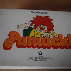 Könnte beinhalten: Eine weiße Box mit einer rothaarigen Comicfigur namens Pumuckl. Die Box ist mit "Babybesteck" und "creation AUERHAHN" beschriftet, mit dem Text "Silberwaren de Tradition".