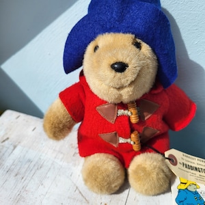 Könnte beinhalten: Ein Plüschtier Paddington Bär, das einen roten Mantel mit braunen Knöpfen und einen blauen Hut trägt. Der Bär sitzt auf einer weißen Holzoberfläche. Das Etikett auf dem Bären lautet "The Paddington Bear Company."