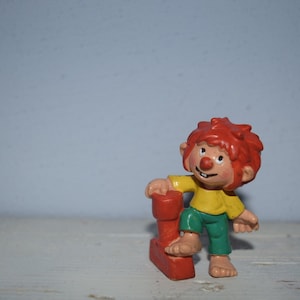 Könnte beinhalten: Eine Figur mit roten Haaren, einem gelben Shirt und grünen Hosen steht neben einem roten und orangen Block. Die Figur hat eine große, rote Nase und ein freundliches Lächeln.