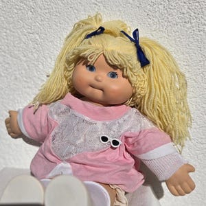 Pode incluir: Uma boneca de pele clara, olhos azuis e longos cabelos de lã amarela presos em duas marias-chiquinhas com fitas azul-escuras. A boneca veste uma camisa rosa com um painel frontal de renda branca, punhos brancos e um pequeno broche de óculos.