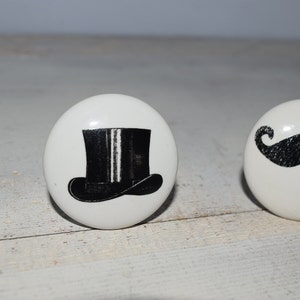 Könnte beinhalten: Zwei weiße Keramikknöpfe mit schwarzen und weißen Designs. Ein Knopf zeigt einen schwarzen Zylinder mit einem weißen Streifen. Der andere Knopf zeigt einen schwarzen Schnurrbart.