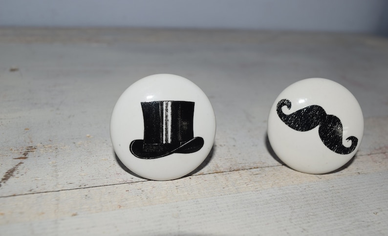 May include: Two white ceramic knobs with black and white designs. One knob features a black top hat with a white stripe. The other knob features a black mustache.