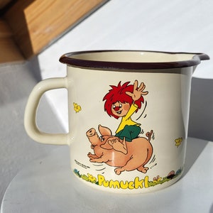 May include: A white enamel mug with a brown rim features a cartoon illustration of a red-haired character riding a pink pig. The character is wearing a yellow shirt and green pants. The mug has the word "Pumuckl" printed on the bottom.