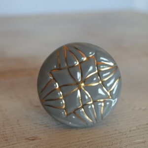 Può includere: Pomo in ceramica grigia con un design geometrico dorato. Il pomo è rotondo e ha una superficie liscia.