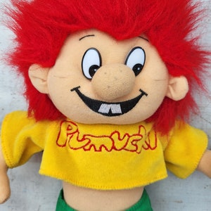 Könnte beinhalten: Ein rothaariges Plüschtier mit einem breiten Grinsen und einem gelben Shirt, auf dem "Pumuckl" steht.