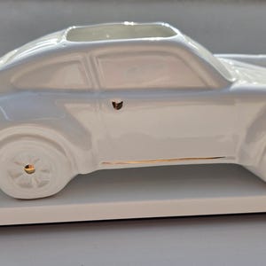 Può includere: Un vaso in ceramica bianca a forma di auto con finitura lucida. Il design del veicolo presenta accenti color oro sulle ruote, sulla maniglia della portiera e lungo la modanatura laterale inferiore. La parte superiore ha un'apertura rettangolare.
