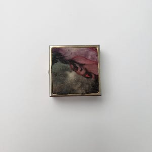 Pode incluir: Uma pequena caixa retangular com uma moldura prateada e um design abstrato colorido no seu interior. O design apresenta tons de rosa, roxo, cinza e preto.