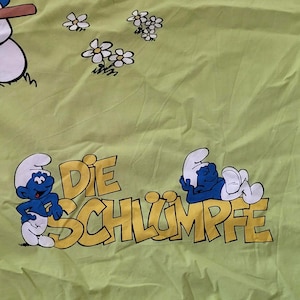 Puede incluir: Tela verde claro con dibujos animados de los Pitufos y el texto "DIE SCHLÜMPFE" en amarillo. Los Pitufos son azules con gorros blancos y están en varias poses, con flores blancas esparcidas por la tela.