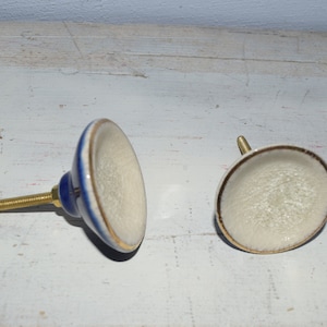 Könnte beinhalten: Zwei Keramik-Schrankknöpfe mit weißer Basis und goldfarbenem Rand. Ein Knopf hat ein blaues Band um die Basis. Beide Knöpfe haben eine Schraube aus Messing.