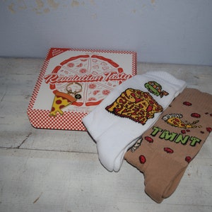 Op de afbeelding: Een set van twee paar sokken met pizza- en Teenage Mutant Ninja Turtles-ontwerpen. De sokken zijn verpakt in een rood-witte pizzadoos met de tekst "Revolution Tasty" erop gedrukt. Een pizzasnij-sleutelhanger is ook inbegrepen.