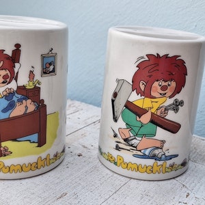 Puede incluir: Dos alcancías de cerámica blanca con un personaje de dibujos animados llamado Pumuckl. Una alcancía muestra a Pumuckl durmiendo en la cama, mientras que la otra lo muestra sosteniendo un martillo y clavos. Ambas alcancías tienen el nombre "Pumuckl" escrito en la parte inferior.