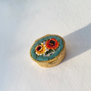Op de afbeelding: Een klein, ovaalvormig sieradendoosje met een bloemenmotief. Het doosje is gemaakt van metaal en heeft een blauw en groen mozaïekpatroon met rode, oranje en gele bloemen.