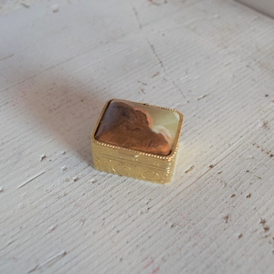 Può includere: Una piccola scatola per gioielli quadrata di colore dorato con un coperchio incernierato. Il coperchio è decorato con un motivo a marmo marrone e verde.