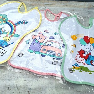 Könnte beinhalten: Drei weiße Baby-Lätzchen mit bunten Comic-Motiven. Das erste Lätzchen hat einen Regenbogen, einen Pinsel und ein Kaninchen. Das zweite Lätzchen hat ein Auto und einen Bären. Das dritte Lätzchen hat einen Bären mit Ballons und einem grünen Rand.