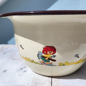 Könnte beinhalten: Eine weiße Emaille-Schüssel mit braunem Rand und einer Comicfigur, die einen Hammer hält. Die Schüssel hat eine Grafik einer Comicfigur mit roten Haaren und einem gelben Hemd, die einen Hammer hält. Die Schüssel hat den Text "Pumuckl" an der Seite.