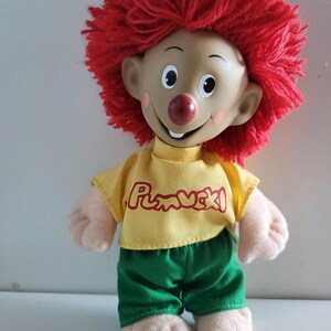 Op de afbeelding: Vintage pop met een lachend gezicht, rood garen haar en een geel shirt met het woord "Pumuckl" erop gedrukt. De pop draagt een groene korte broek en heeft beige handen en voeten.