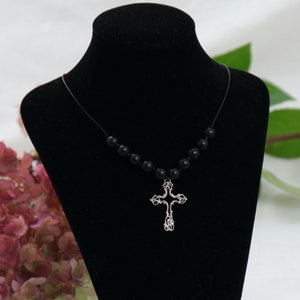 Könnte beinhalten: Eine schwarze Halskette mit schwarzen Perlen und einem silbernen Kreuzanhänger auf einem schwarzen Samt-Präsentationsständer.