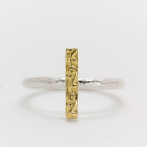Könnte beinhalten: Ein silberner Ring mit einem goldenen Balken in der Mitte. Der goldene Balken hat ein dekoratives Muster.