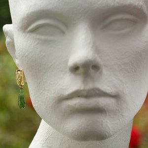Peut inclure: Un mannequin blanc portant une paire de boucles d'oreilles en or avec des gouttes de pierres pr&eacute;cieuses vertes.