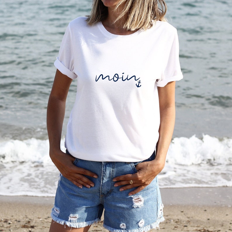 Puede incluir: Camiseta blanca con la palabra "moin" y un ancla impresa en azul.