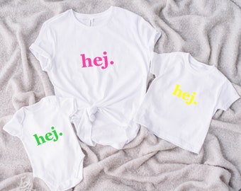 Puede incluir: Una camiseta blanca de manga corta con el estampado "hej." en rosa, una camiseta blanca más pequeña de manga corta con el estampado "hej." en amarillo y un body blanco de manga corta con el estampado "hej." en verde, todos con cuello redondo.