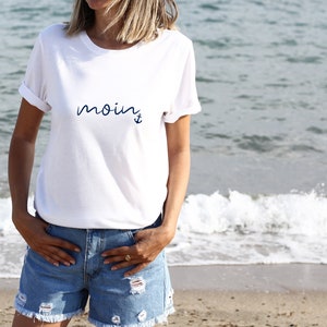 Puede incluir: Camiseta blanca con la palabra "moin" y un ancla impresa en azul.