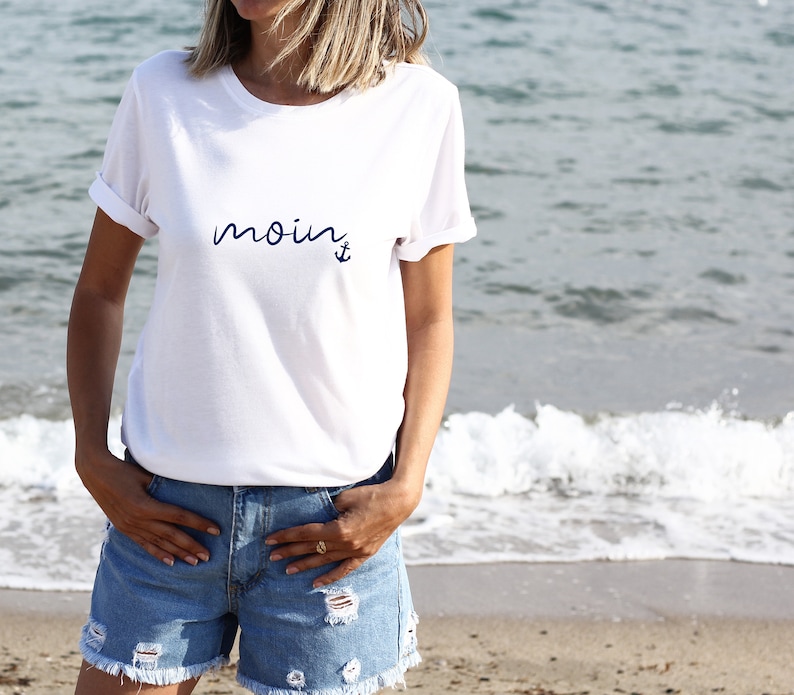 Puede incluir: Camiseta blanca con la palabra "moin" y un ancla impresa en azul.