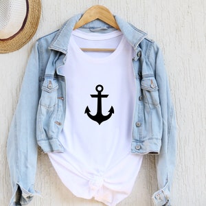 Könnte beinhalten: Ein weißes T-Shirt mit einem schwarzen Anker-Motiv. Das Shirt hängt an einem Kleiderbügel und wird über einer Jeansjacke getragen.